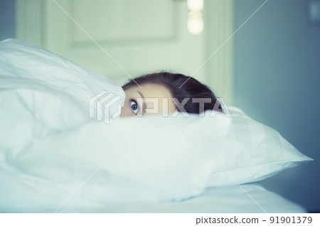 A young woman with huge frightened open eyes lying in bed and covers herself with the sheets and hides. Nightmares, insomnia, phobias, panic attacks. Concept A young woman with huge frightened open eyes lying in bed and covers herself with the sheets and hides. Nightmares, insomnia, phobias, panic attacks. Concept 91901379