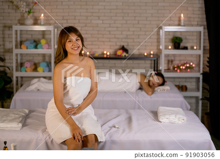 Wide shot of Asian beautiful woman wearing towel look at camera and sit on bed in front of other woman lie on bed during spa and massage for good health. 91903056