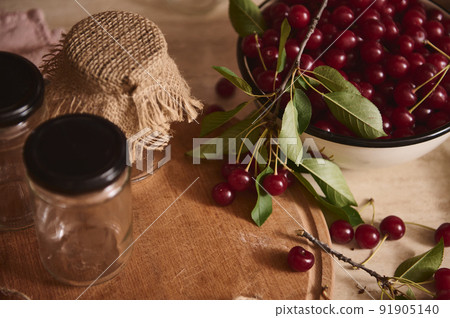 Top view of sterile jars with lids on wooden board next to a bowl with freshly picked cherries on the kitchen countertop 91905140