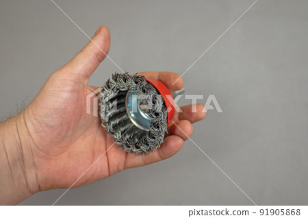 A man's hand holds a bowl wire brush. A tool for woodworking as well as stripping surfaces of corrosion, scale, or rust. Side view. Gray background. Selective focus. 91905868