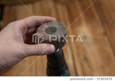 Man places flap wheel in cam of an electric drill chuck. Tool for woodworking, sanding, removing rust from various surfaces. Yellow wooden boards defocused in background. Close-up. Selective focus. 91905870
