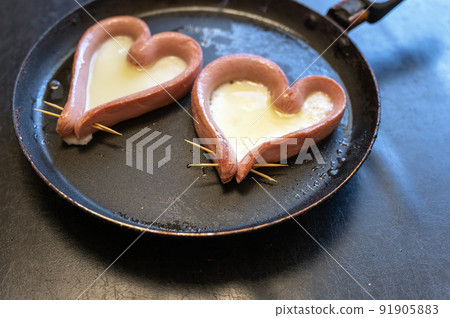 Two heart-shaped sausages lie in the pan. Roasting sausages cut lengthwise. Thick albumen poured inside. Cooking a meal. Love, relationship concept. Selective focus. Two heart-shaped sausages lie in the pan. Roasting sausages cut lengthwise. Thick albumen poured inside. Cooking a meal. Love, relationship concept. Selective focus. 91905883