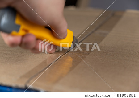 A man's hand opens a cardboard box with a yellow cutter. The flaps of the package are sealed with adhesive tape. Close-up. Selective focus. 91905891
