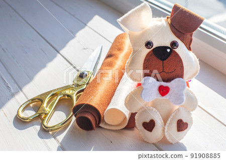 A beautiful puppy made of felt with a brown ear and muzzle, with a bone and a red heart sits on a background of white boards. Tailor's scissors, felt rolls. Creativity, hobby, sewing toys with a child 91908885
