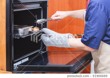 A chef in an apron and protective gloves pulls meat medallions out of the oven with meat tongs. Kitchen, food preparation, cooking. Restaurant, cafe, eatery. Cooking courses, school. A chef in an apron and protective gloves pulls meat medallions out of the oven with meat tongs. Kitchen, food preparation, cooking. Restaurant, cafe, eatery. Cooking courses, school. 91908889