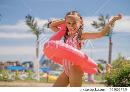 A beautiful girl holds a flamingo-shaped lifebuoy against the backdrop of a resort with palm trees and blue sky. Tourist holidays 91909789