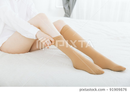Beautiful long female legs in stockings. Girl putting on stockings at home in a white room. Beige knee socks. Varicose veins prevention. Woman body in underwear. 91909876
