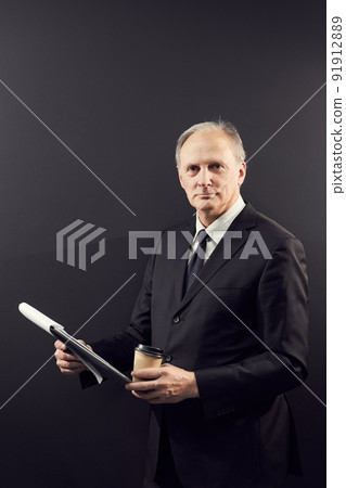 Portrait of content confident owner of business company standing against dark wall and analyzing report in clipboard while drinking coffee 91912889