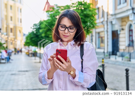 Middle aged smiling woman looking into smartphone on city street 91913134