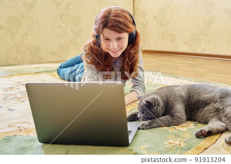 Preteen girl in headphones and gray british cat looking at laptop together Preteen girl in headphones and gray british cat looking at laptop together 91913204