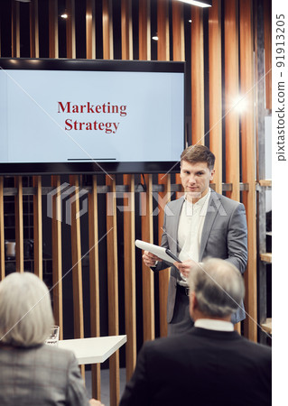 Content young social media manager in gray suit standing in front of audience and holding clipboard while giving speech at conference Content young social media manager in gray suit standing in front of audience and holding clipboard while giving speech at conference 91913205