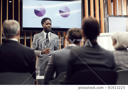 Content confident young Afro-American financial specialist in gray suit standing against plasma screen with graph charts and giving speech in front of business colleagues Content confident young Afro-American financial specialist in gray suit standing against plasma screen with graph charts and giving speech in front of business colleagues 91913223