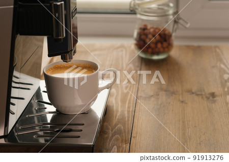 Automatic coffee machine for coffee in white cup near window at daytime in a coffee shop, cafe or restaurant. Jar of hazelnuts on background 91913276