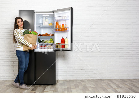 Young woman with paper bag full of products near open refrigerator indoors, space for text Young woman with paper bag full of products near open refrigerator indoors, space for text 91915556
