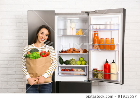 Young woman with paper bag full of products near open refrigerator indoors 91915557