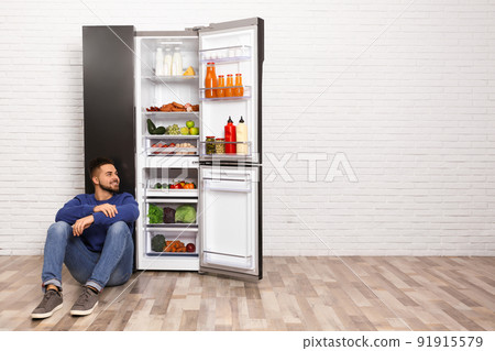 Happy young man sitting on floor near refrigerator indoors, space for text Happy young man sitting on floor near refrigerator indoors, space for text 91915579
