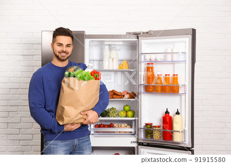 Young man with paper bag full of products near open refrigerator indoors 91915580