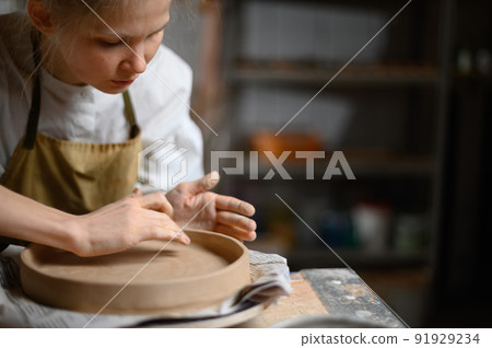 A ceramist makes a plate. Woman in an apron works in a pottery workshop. A ceramist makes a plate. Woman in an apron works in a pottery workshop. 91929234