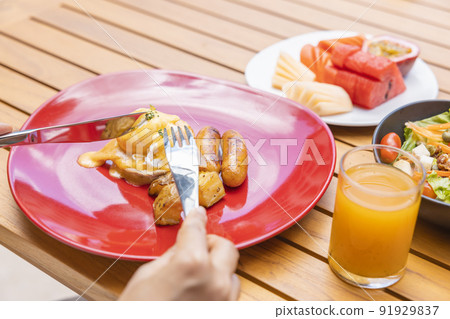 Woman's hand holding a fork and eating Breakfast. Egg Benedict, fruits such as watermelon, papaya, melon, passion fruit, orange juice and coffee. placed on a gray placemat Woman's hand holding a fork and eating Breakfast. Egg Benedict, fruits such as watermelon, papaya, melon, passion fruit, orange juice and coffee. placed on a gray placemat 91929837