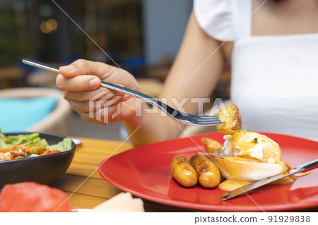 Woman's hand holding a fork and eating Breakfast. Egg Benedict, fruits such as watermelon, papaya, melon, passion fruit, orange juice and coffee. placed on a gray placemat Woman's hand holding a fork and eating Breakfast. Egg Benedict, fruits such as watermelon, papaya, melon, passion fruit, orange juice and coffee. placed on a gray placemat 91929838