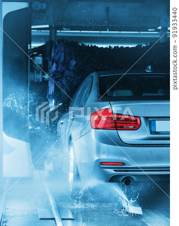 A grey business sedan vehicle running through an automatic car wash with brushes and water hitting car. A grey business sedan vehicle running through an automatic car wash with brushes and water hitting car. 91933440