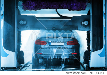 A grey business sedan vehicle running through an automatic car wash with brushes and water hitting car. 91933441