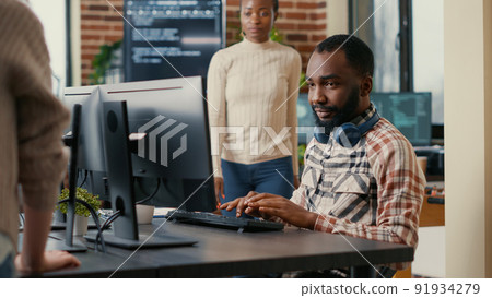 Relaxed software developer wearing wireless headphones smiling working looking at computer screen while typing. Coder writing code in it agency office with colleague asking for informations. 91934279