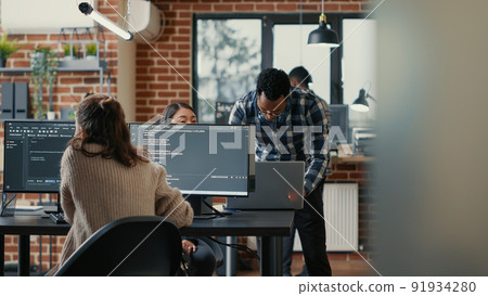 Database programer writing code in front of multiple computer screens displaying artificial intelligence algorithm. Developer coding database while colleagues doing teamwork in background. 91934280