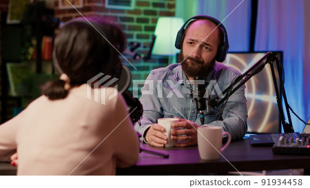 Man and woman having casual conversation and drinking coffe or tea in online podcast using professional recording equipment in home studio. Radio show host broadcasting interview with influencer. Man and woman having casual conversation and drinking coffe or tea in online podcast using professional recording equipment in home studio. Radio show host broadcasting interview with influencer. 91934458