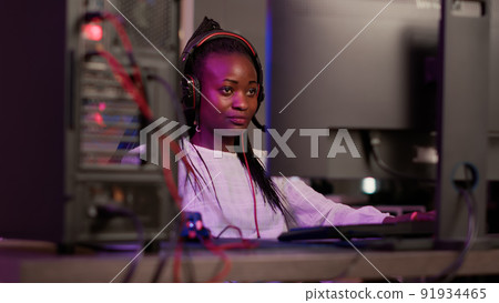 Handheld shot of african american streamer talking on headset while online playing first person shooter on gaming pc setup. Gamer girl streaming while sitting in gaming chair explaining gameplay. 91934465