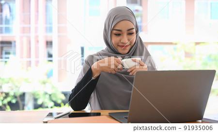 Pleased muslim lady in hijab holding cup of coffee and reading business email or financial information on laptop Pleased muslim lady in hijab holding cup of coffee and reading business email or financial information on laptop 91935006