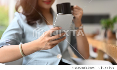Relaxed female entrepreneur drinking coffee and checking online news or email on her smart phone 91935195