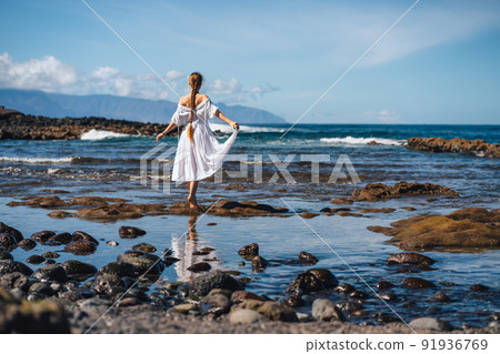 Back view young happiness traveller woman in white dress standing on rocky volcanic beach. Cute girl enjoy tropical sea on relax holiday vacation summer sunny day. Tenerife, Canary Islands, Spain 91936769