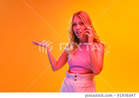 Portrait of young beautiful woman posing isolated over yellow background in neon light. Expressive phone talk 91936847