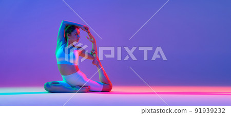 Studio shot of young flexible girl in fitness sport uniform practicing isolated on gradient pink-purple background in neon light. Sport, beauty, ad Studio shot of young flexible girl in fitness sport uniform practicing isolated on gradient pink-purple background in neon light. Sport, beauty, ad 91939232