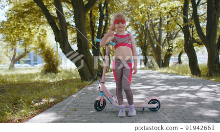Athletic fit young girl rides on electric scooter, standing near bike looking at camera in park 91942661