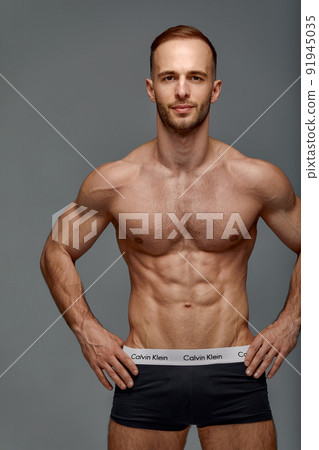 Portrait of a handsome young guy model a shirtless with a muscular healthy body, showing off his biceps muscles on a gray background 91945035