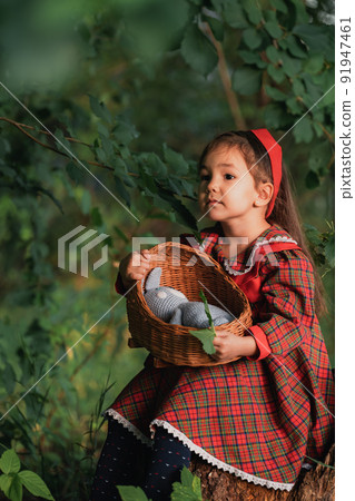 Little girl in red dress holding wicker cradle with doll in front of nusto forest Little girl in red dress holding wicker cradle with doll in front of nusto forest 91947461