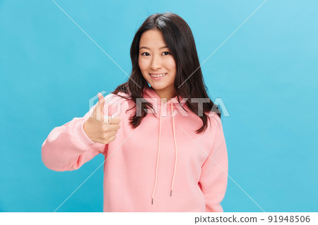 Friendly cute Asian student young lady in pink hoodie sweatshirt show thumb up gesture Like posing isolated on over blue studio background. The best offer for ad. People Emotions for Everyday concept 91948506