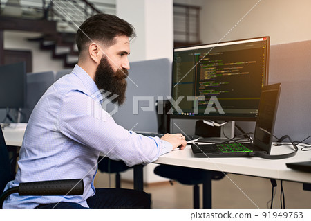 Software programming. Bearded man working on computer in IT office, sitting at desk writing code, working on a project in software development company or startup. High quality image. 91949763