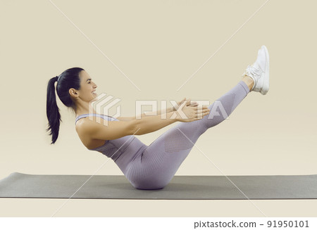 Smiling sporty woman in sportswear doing abs exercise with leg raise on beige background. 91950101