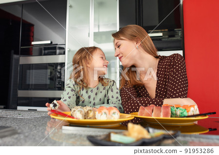 Beautiful caucasian mom and daughter eating rolls and sushi at family lunch at home. Delivery food. Traditional japanese food. Beautiful caucasian mom and daughter eating rolls and sushi at family lunch at home. Delivery food. Traditional japanese food. 91953286