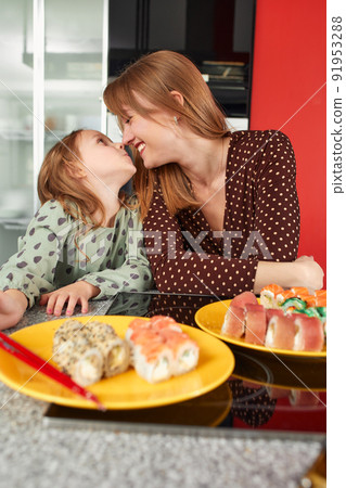 Beautiful caucasian mom and daughter eating rolls and sushi at family lunch at home. Delivery food. Traditional japanese food. Beautiful caucasian mom and daughter eating rolls and sushi at family lunch at home. Delivery food. Traditional japanese food. 91953288