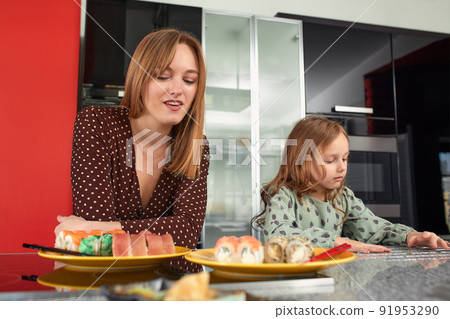Beautiful caucasian mom and daughter eating rolls and sushi at family lunch at home. Delivery food. Traditional japanese food. Beautiful caucasian mom and daughter eating rolls and sushi at family lunch at home. Delivery food. Traditional japanese food. 91953290