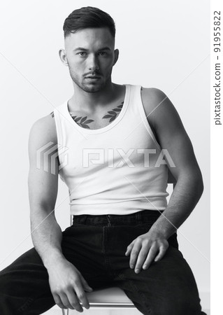 Modelling snapshots. Serious self-confident attractive handsome man in basic white undershirt sitting on chair posing in white studio background. Black and White concept. Copy space 91955822