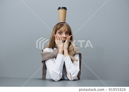 Excited young blonde student lady in warm sweater with takeaway coffee on head touch cheeks open mouth sitting in gray modern home office. Coffee break Lover Concept. Copy space. Cool offer Excited young blonde student lady in warm sweater with takeaway coffee on head touch cheeks open mouth sitting in gray modern home office. Coffee break Lover Concept. Copy space. Cool offer 91955826