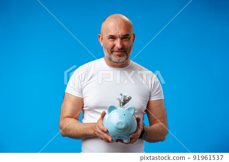 Middle-aged man holding piggy bank against blue background 91961537