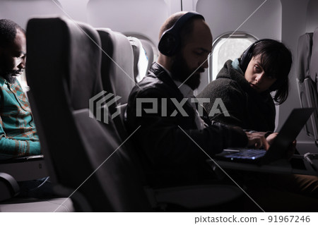 Female passenger looking at man during airplane flight, using laptop online on aerial transportation. Group of tourists travelling by plane on international airline service, aviation jet. 91967246