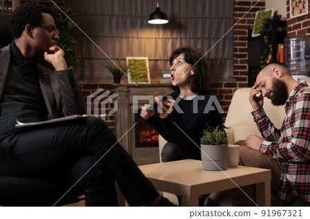 Unhappy wife showing husband proof of cheating and adultery on smartphone at couple therapy session. Marriage betrayal and crisis with suspicious text messages, wanting to divorce. Unhappy wife showing husband proof of cheating and adultery on smartphone at couple therapy session. Marriage betrayal and crisis with suspicious text messages, wanting to divorce. 91967321