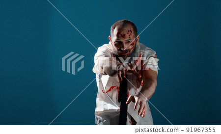 Growling creepy monster prowling at camera on blue background. Mindless looking apocalyptic brain-eating evil monster with deep and bloody wounds acting bizarre and sinister. Studio shot Growling creepy monster prowling at camera on blue background. Mindless looking apocalyptic brain-eating evil monster with deep and bloody wounds acting bizarre and sinister. Studio shot 91967355